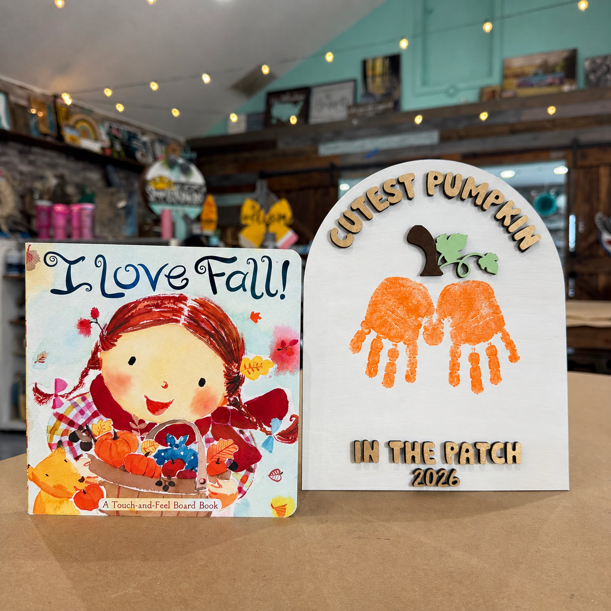Two autumn-themed products displayed side-by-side on a wooden table in a shop: a children's board book titled 'I Love Fall!' and a personalized wooden sign that reads 'CUTEST PUMPKIN IN THE PATCH 2026' with orange handprint pumpkin designs.