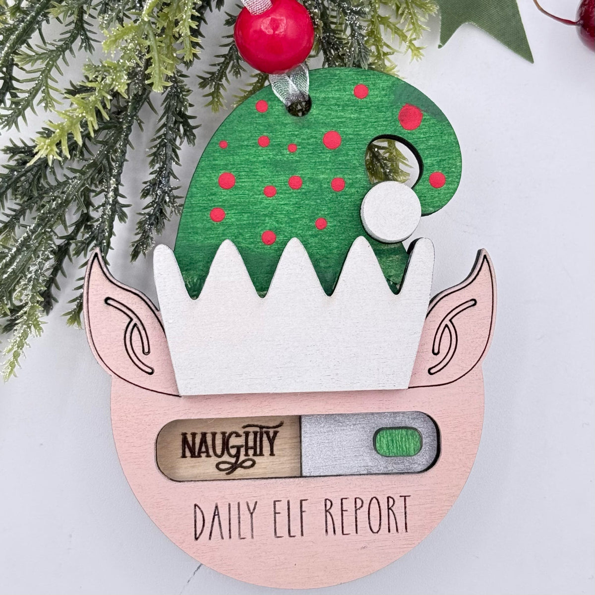 A wooden Christmas ornament shaped like an elf's head with a green hat, pink cheeks, and a progress-bar style window labeled 'NAUGHTY DAILY ELF REPORT'. Green pine branches and red ornaments decorate the top against a white background.