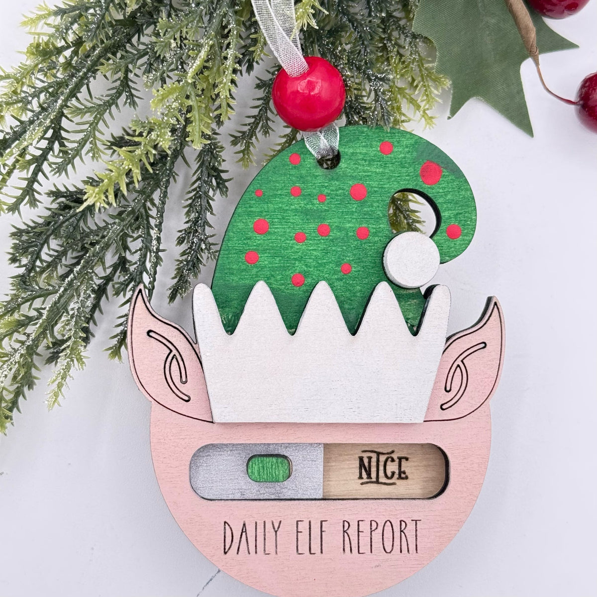 A festive wooden Christmas ornament shaped like an elf's head, hanging with a red berry ornament and ribbon. The ornament has a green hat with red dots, a white beard, pink ears, and a lower section labeled 'DAILY ELF REPORT' with a small window showing a green progress tab and a wooden 'NICE' tab.
