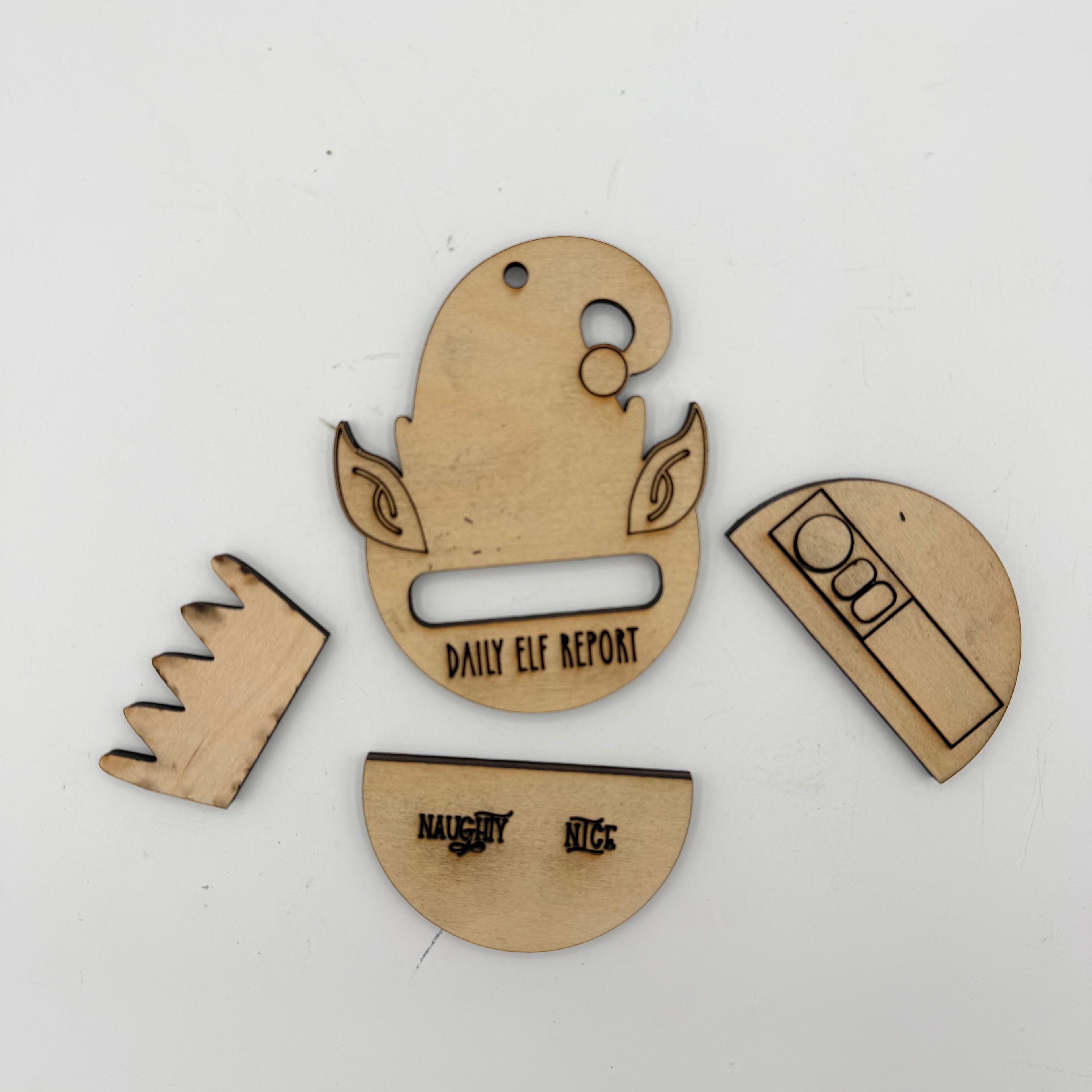Four laser-cut wooden pieces for a daily elf report tracker, including a half-circle with a slot, a crown, a half-circle with text, and a semi-circle with icons, shown on a white background.