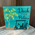 A lighted sign with a blue background featuring illustrations of palm trees and a cartoon golf cart with the text 'DECK THE PALMS'.