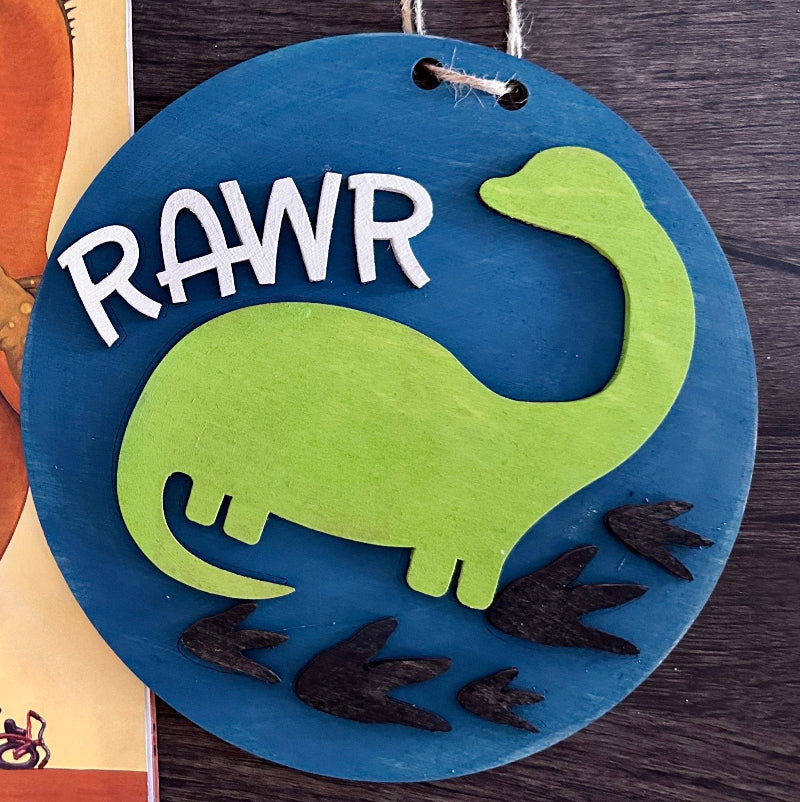 A round decorative plaque with a green dinosaur and the word 'RAWR' on it, placed against a backdrop of a children's book.