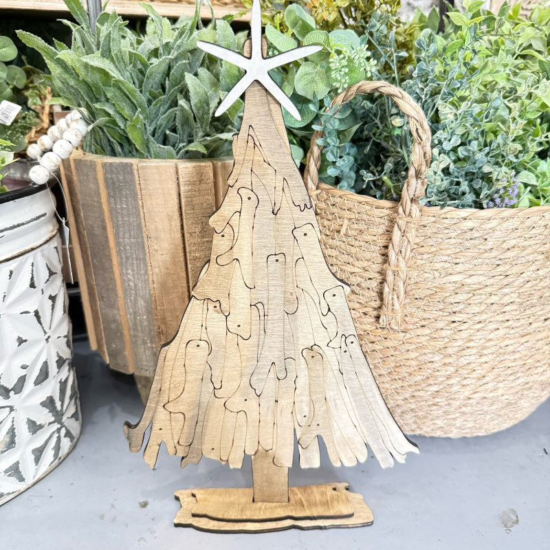 A rustic wooden Christmas tree decoration with a star topper, displayed in a home decor setting alongside potted plants and a woven basket.
