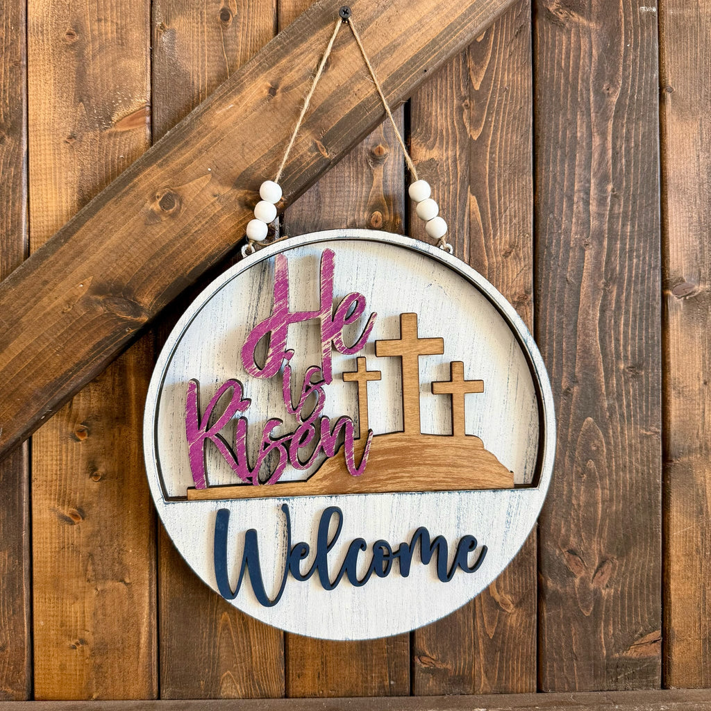 Decorative sign with 'He is Risen' and 'Welcome' text on a wooden background