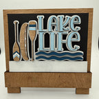 Decorative sign with 'Lake Life' text and oar designs on a black background.