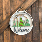 Decorative sign with Christmas trees and 'Welcome' text on a wooden background