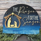 Decorative sign with nativity scene and 'The Reason For the Season' text on a wooden background.