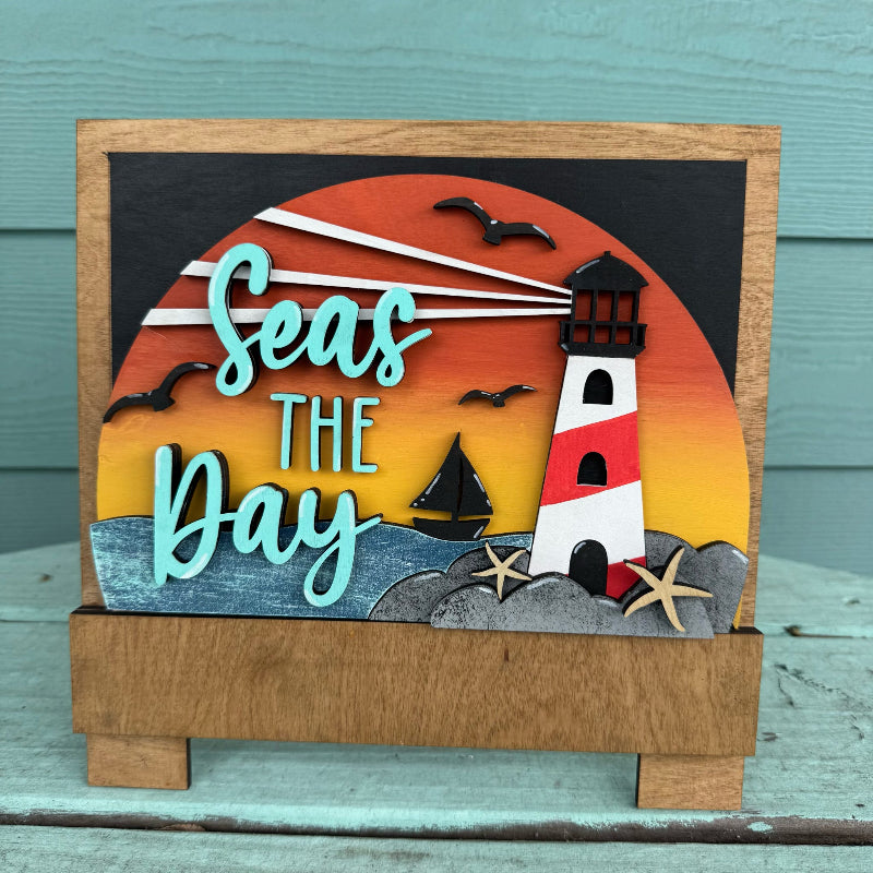 Decorative sign with lighthouse and 'Seas the Day' text on a wooden stand.