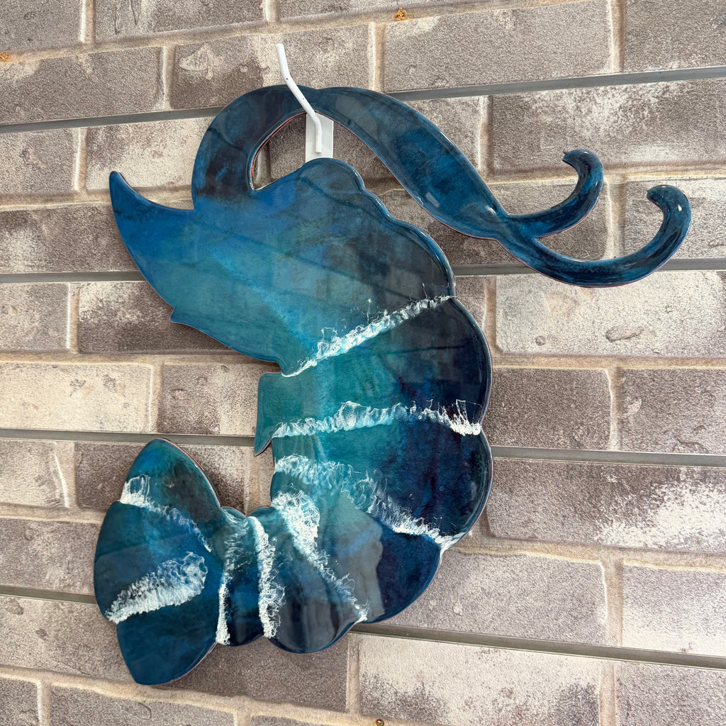 A decorative wall hanging shaped like a stylized fish or wave, made from glossy blue glass with white marbled streaks, photographed against a gray brick wall.