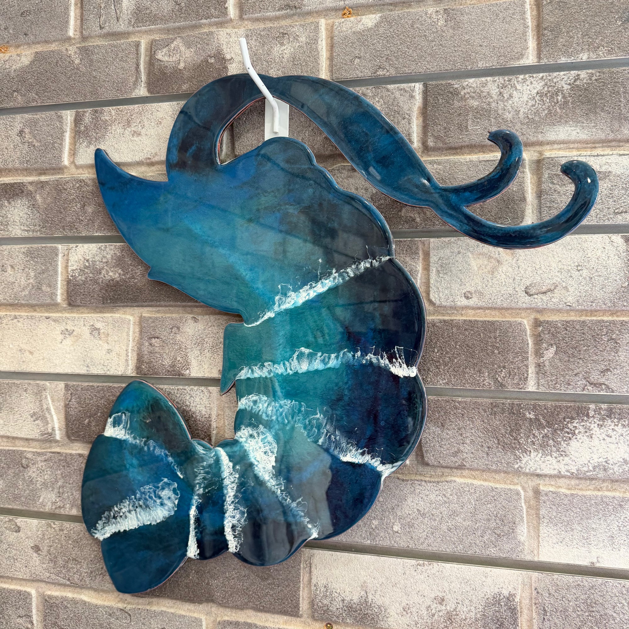 A decorative wall hanging shaped like a stylized fish or wave, made from glossy blue glass with white marbled streaks, photographed against a gray brick wall.