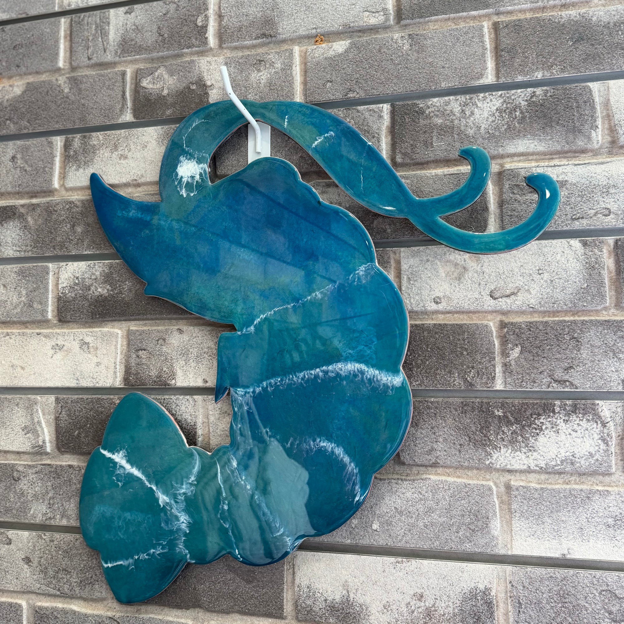 A teal blue glass wall art piece shaped like a stylized fish or shell, mounted against a gray brick wall.