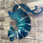A blue and teal glass wall hanging shaped like a stylized wave or shell, with white resin-like streaks and a white hanging loop, mounted against a gray brick wall.