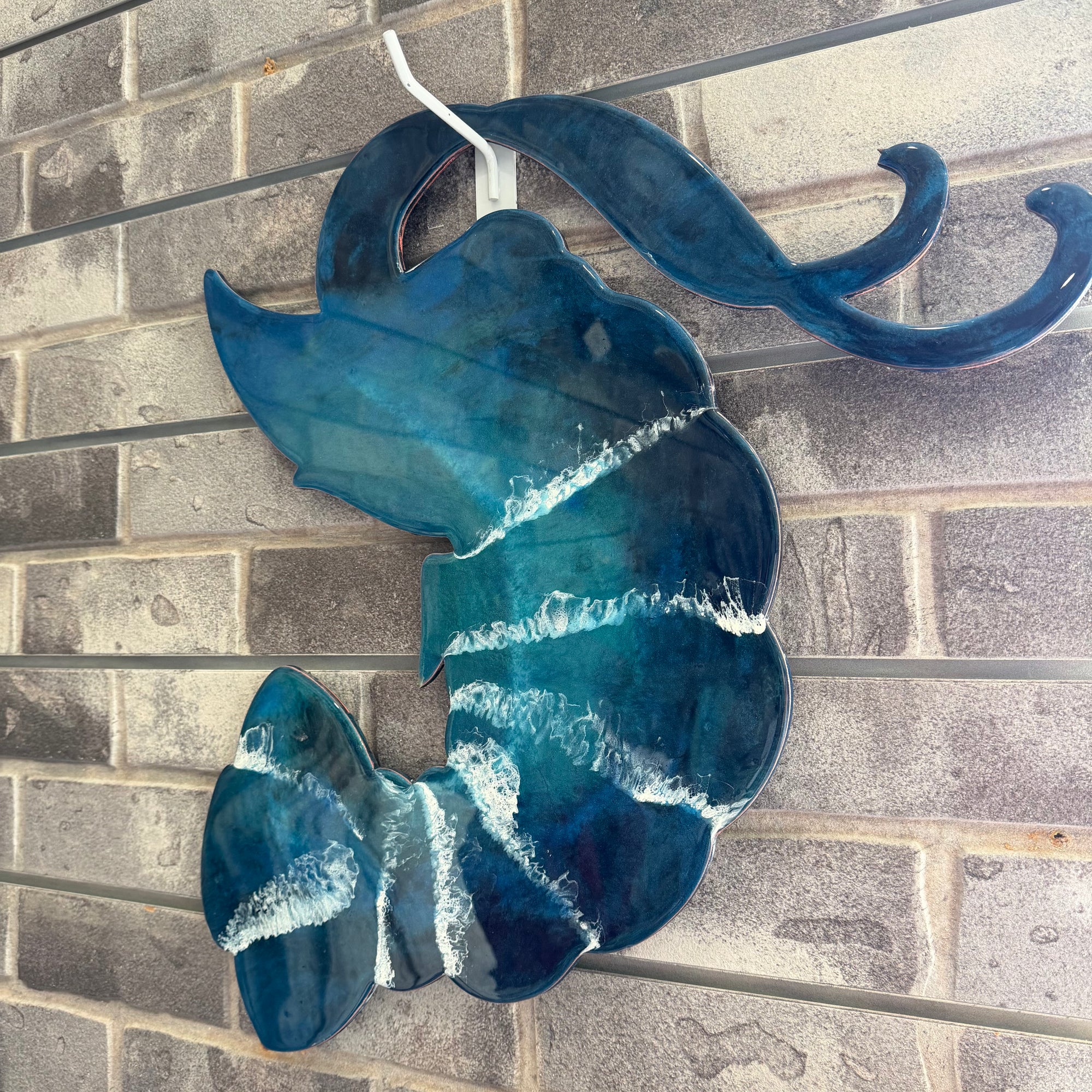 A blue and teal glass wall hanging shaped like a stylized wave or shell, with white resin-like streaks and a white hanging loop, mounted against a gray brick wall.