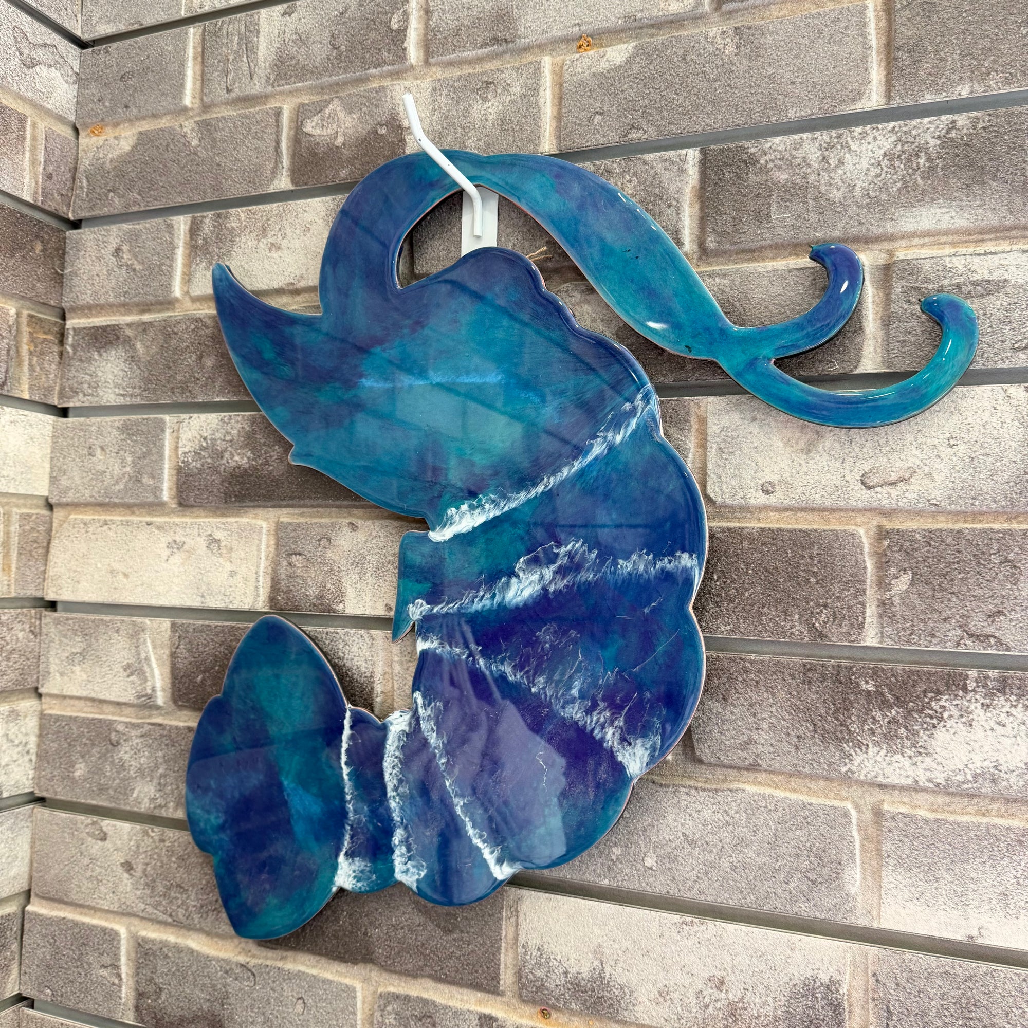 A blue and teal glass art piece shaped like a stylized mermaid tail with white resin-like swirls, mounted on a gray brick wall.