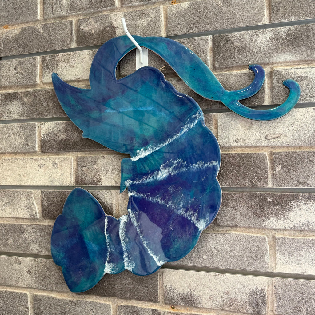 A decorative wall hanging shaped like a stylized fish or abstract marine form, made from glossy blue and purple glass with white marbled veining, displayed against a gray brick wall.