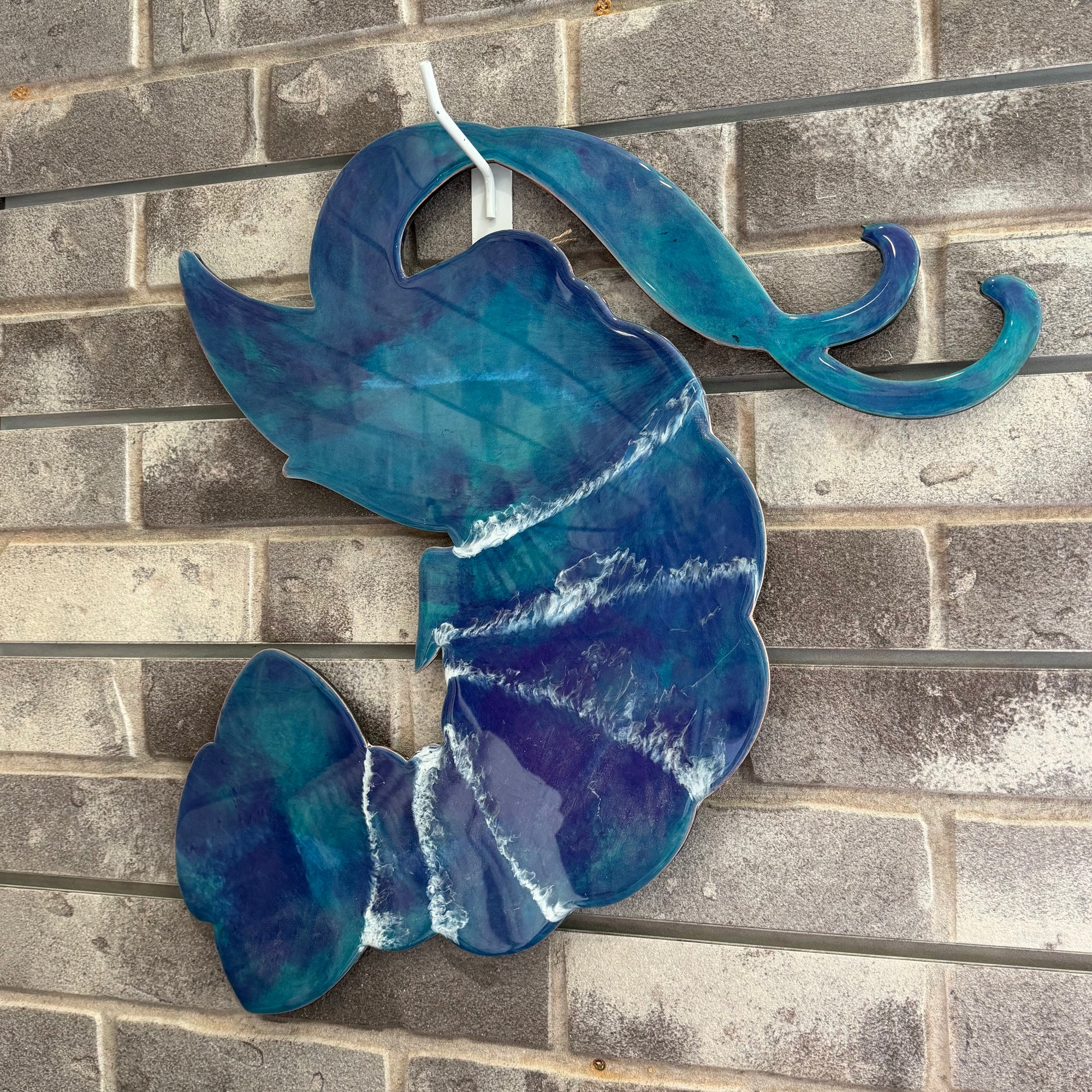 A decorative wall hanging shaped like a stylized fish or abstract marine form, made from glossy blue and purple glass with white marbled veining, displayed against a gray brick wall.