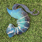 A decorative metal wall hanging shaped like a stylized mermaid tail, featuring a blue and teal galaxy design with white star-like speckles, displayed on green artificial turf.