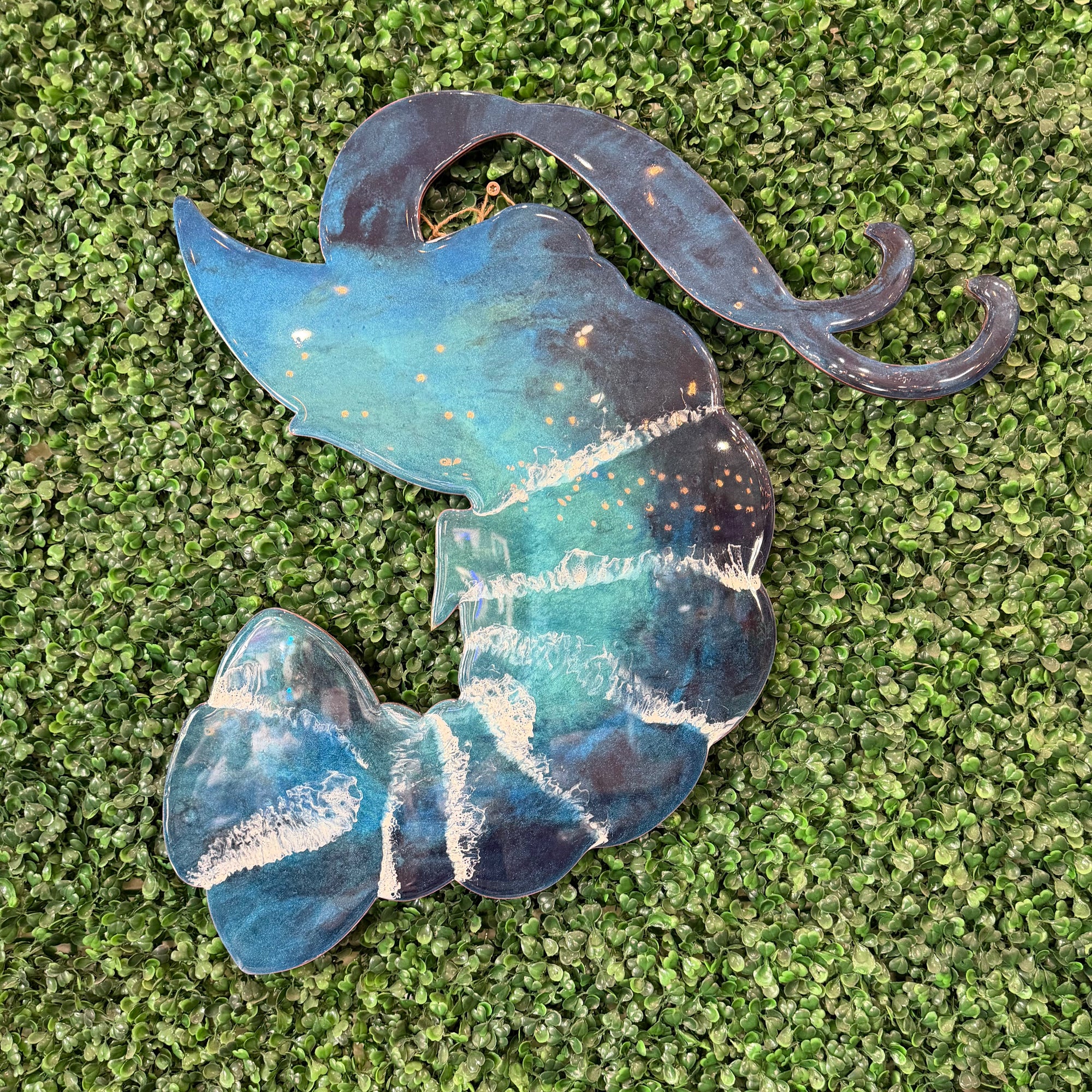 A decorative metal wall hanging shaped like a stylized mermaid tail, featuring a blue and teal galaxy design with white star-like speckles, displayed on green artificial turf.