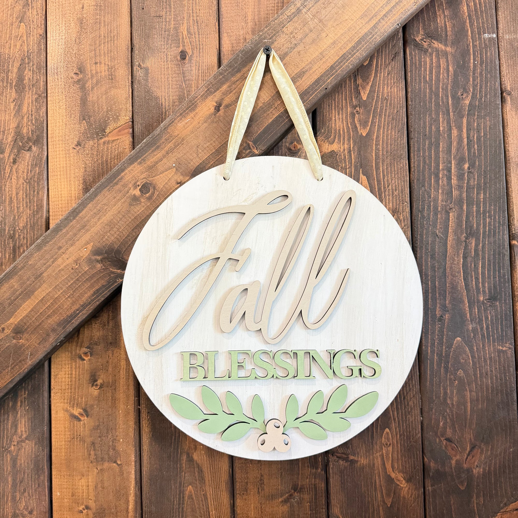 Round wooden hanging sign with the words “Fall Blessings” in light wood and pale green, decorated with green leaves and a small acorn, hanging against a rustic dark wood plank background.