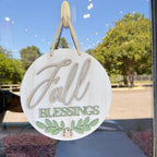Round wooden hanging sign with 'Fall Blessings' text and green leaf design, suspended by twine on a glass door with outdoor trees and sky reflected behind it.