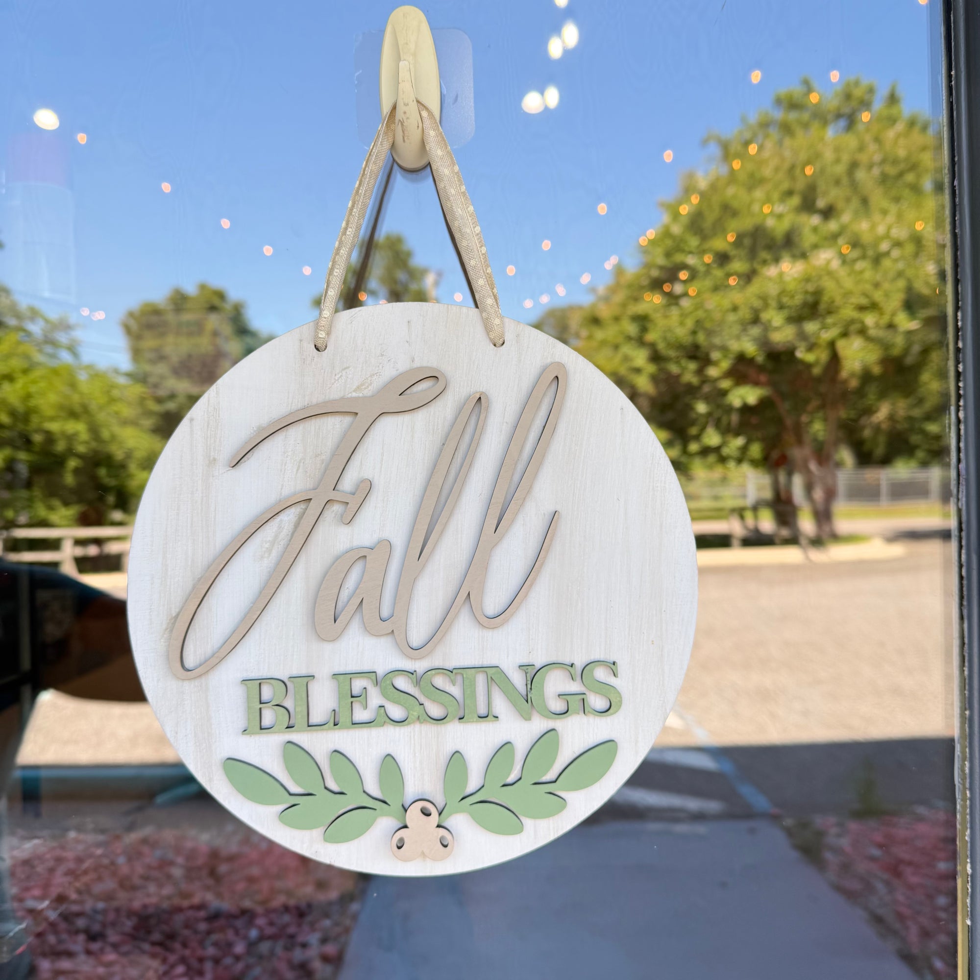 Round wooden hanging sign with 'Fall Blessings' text and green leaf design, suspended by twine on a glass door with outdoor trees and sky reflected behind it.