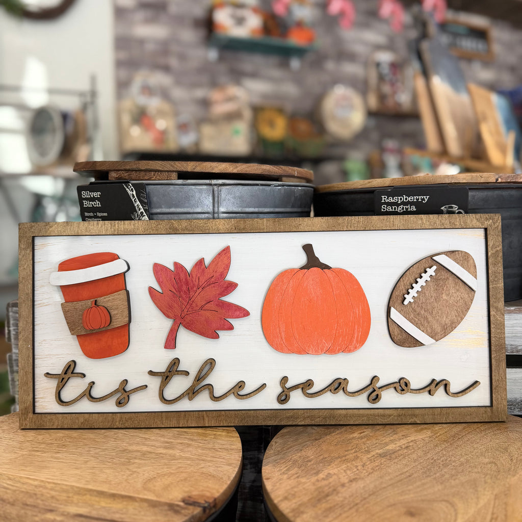 A rectangular wooden sign with a whitewashed face and natural wood edges, featuring autumn-themed illustrations and the phrase "tis the season" in cursive. The design includes an orange coffee cup with a small pumpkin, a red maple leaf, an orange pumpkin, a brown-and-white football, and cursive text.
