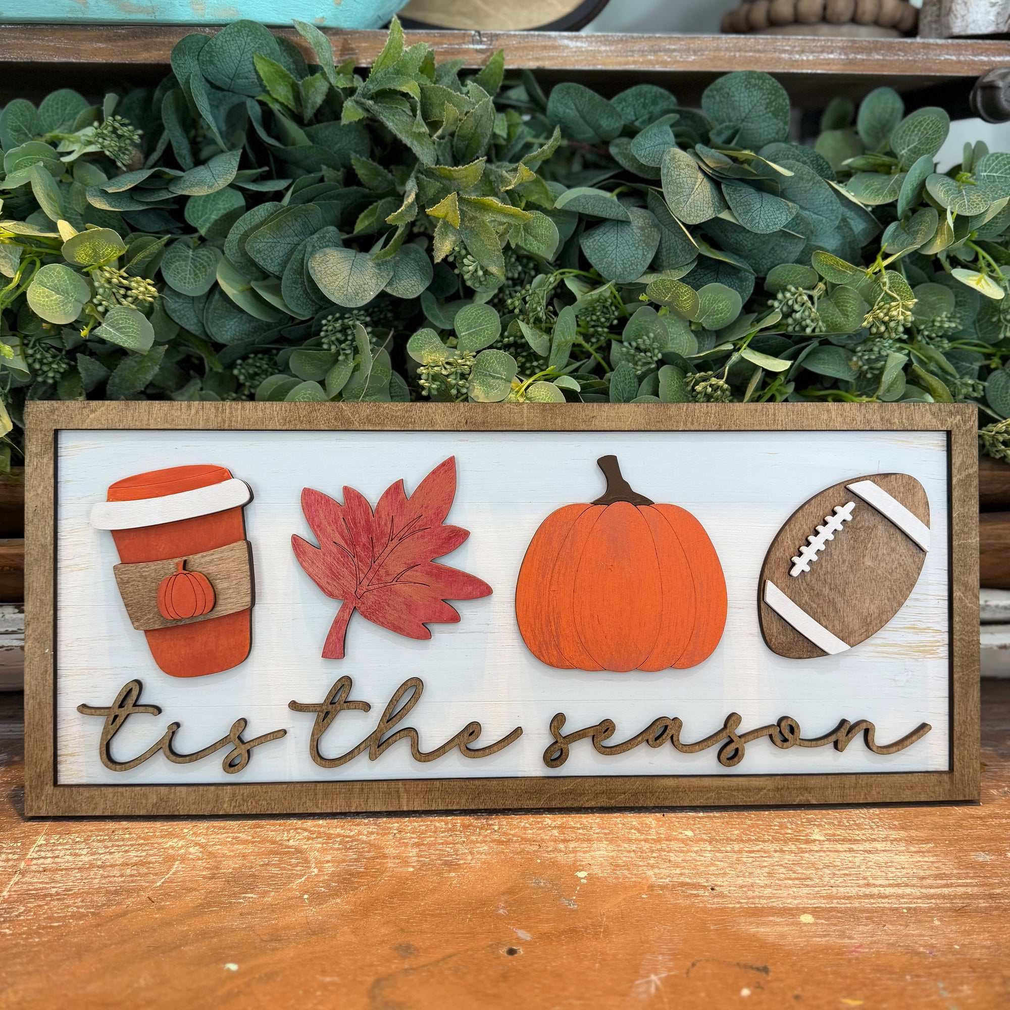 A rectangular wooden sign with a white distressed background and raised autumn-themed cutouts, including a coffee cup with a pumpkin, a red leaf, a pumpkin, a football, and the phrase "tis the season" in script. The sign is displayed on a wooden surface in front of green foliage.