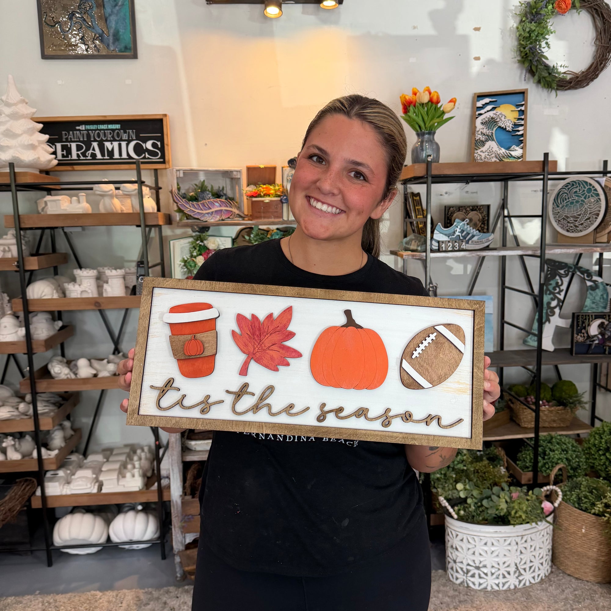 A smiling woman in a black shirt holds up a rustic wooden sign that reads 'tis the season' with autumn-themed illustrations including a coffee cup, maple leaf, pumpkin, and football. The background shows a decorated craft or home decor store with shelves of ceramic items and plants.