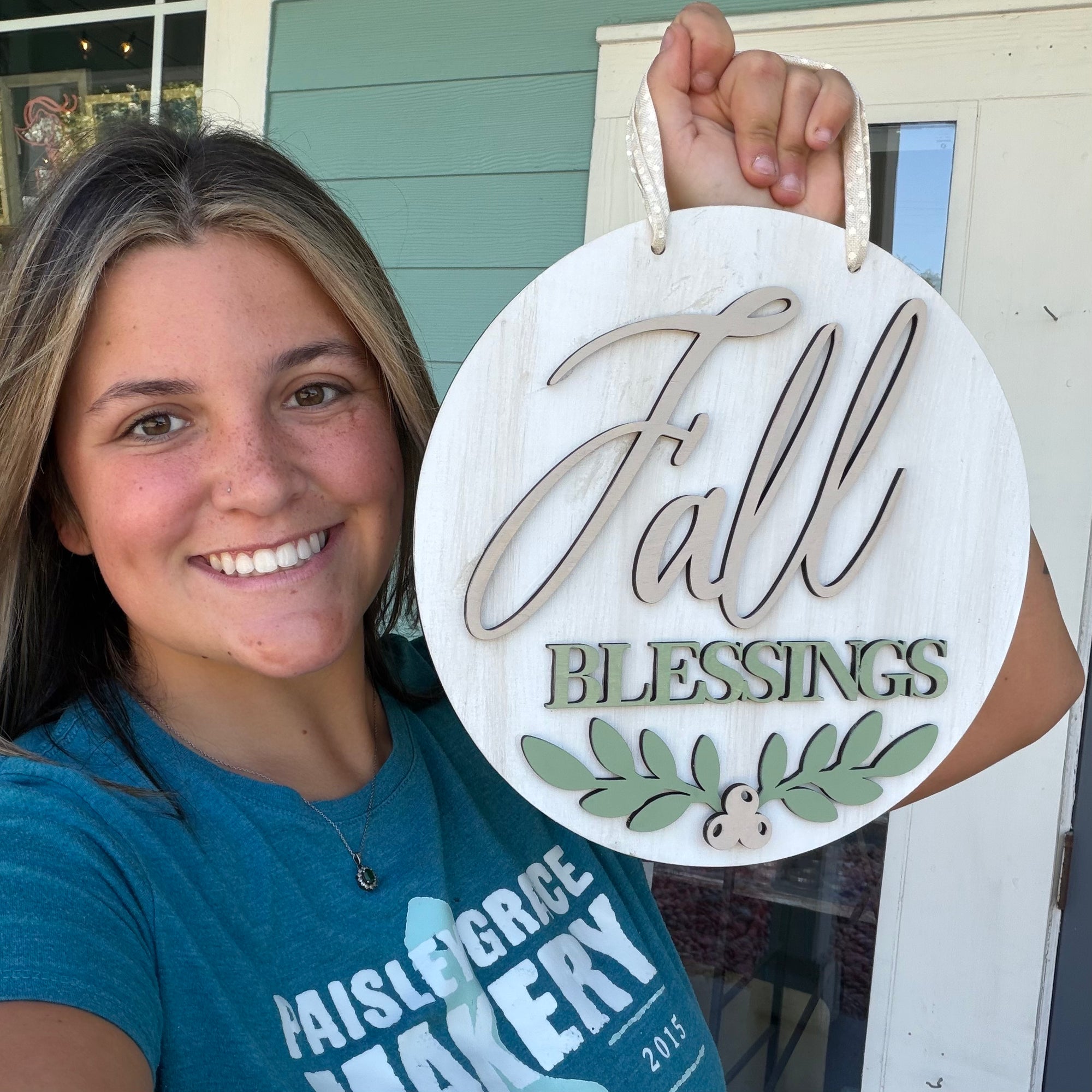 A smiling woman holds up a round white wooden sign that reads 'Fall Blessings' with green leafy accents, photographed outdoors in front of a house door.