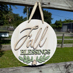 Round wooden hanging sign with the words “Fall Blessings” and a small leaf motif, photographed outdoors against a fence and grassy background.