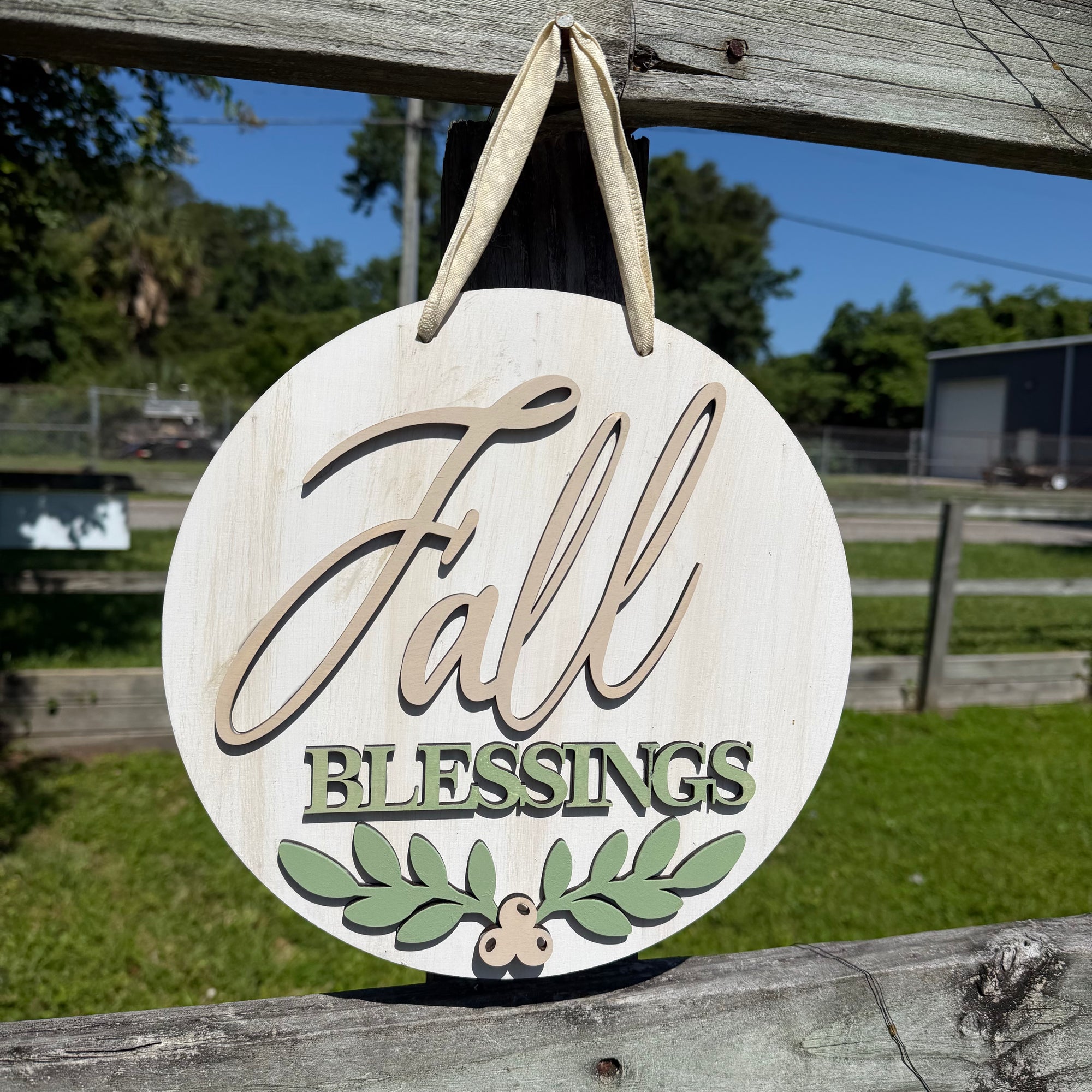 Round wooden hanging sign with the words “Fall Blessings” and a small leaf motif, photographed outdoors against a fence and grassy background.
