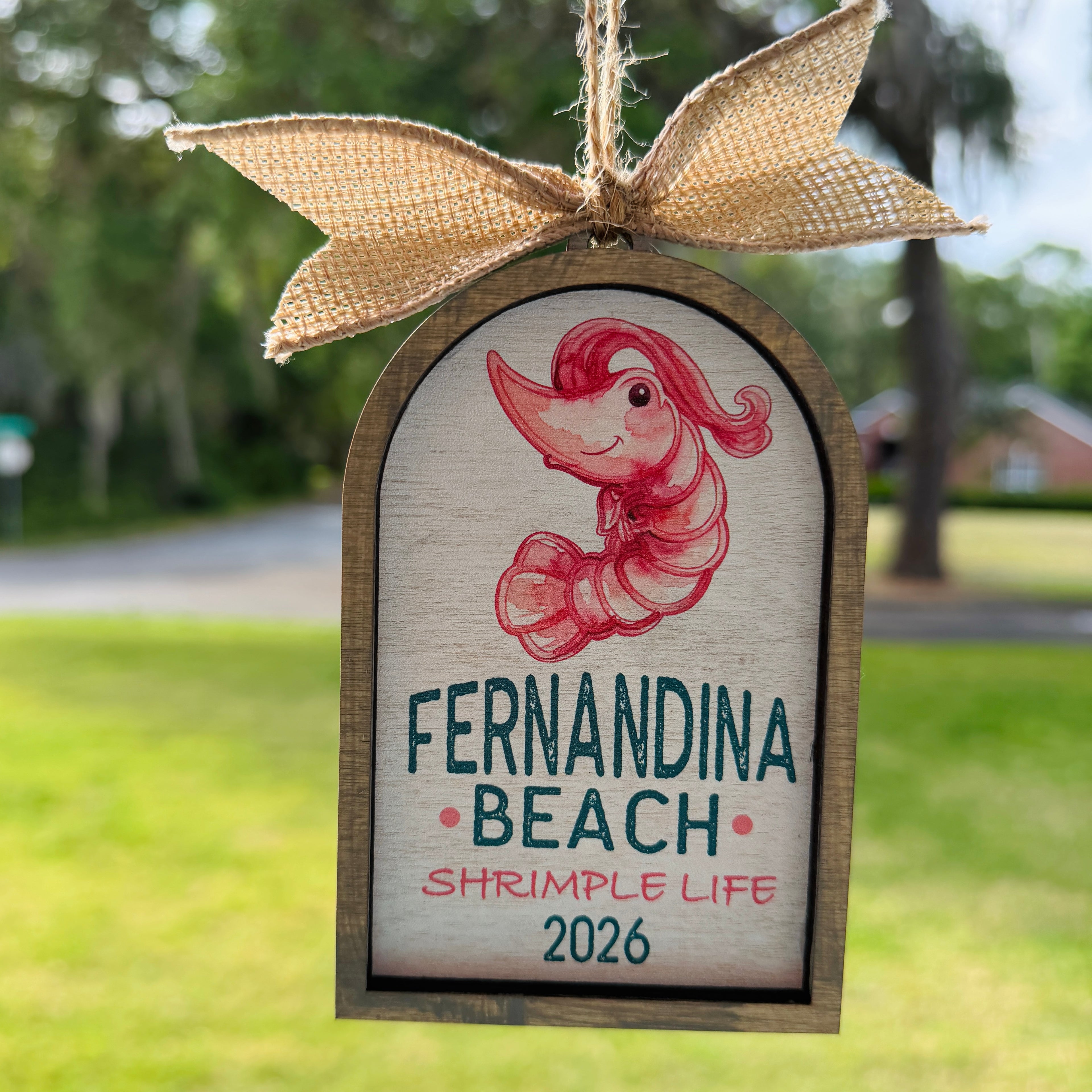 Decorative sign with a shrimp illustration and text 'Fernandina Beach Shrimple Life 2026' on a wooden frame with a burlap bow.