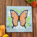 A decorative folk art butterfly painting with vibrant colors and intricate details, displayed on a wooden surface surrounded by floral decorations.