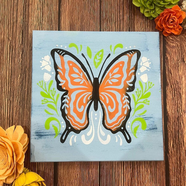 A decorative folk art butterfly painting with vibrant colors and intricate details, displayed on a wooden surface surrounded by floral decorations.