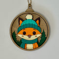 Wooden ornament with a fox wearing a teal hat and scarf on a plain background