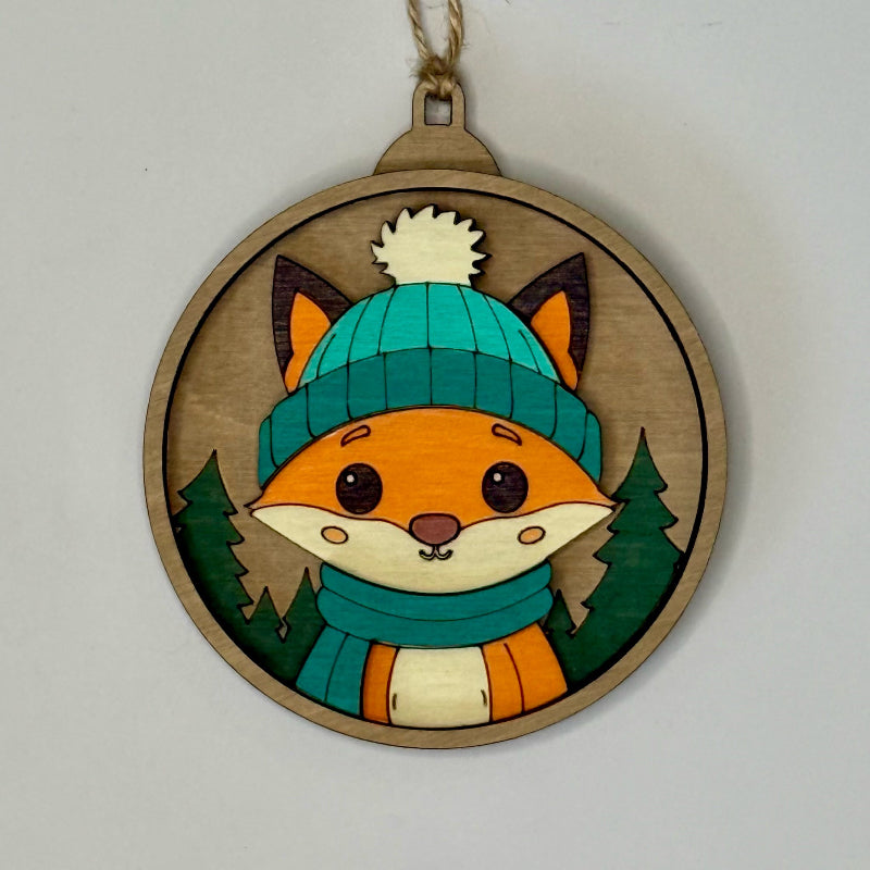 Wooden ornament with a fox wearing a teal hat and scarf on a plain background