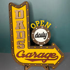A decorative sign with a yellow background and the text 'DADS Garage' with a arrow pointing to the right, and a smaller circle with the text 'OPEN daily'.