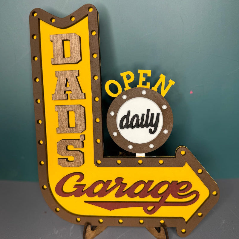 A decorative sign with a yellow background and the text 'DADS Garage' with a arrow pointing to the right, and a smaller circle with the text 'OPEN daily'.
