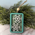 Decorative ornament with snowflake design on a teal background, hanging against a green leafy background.