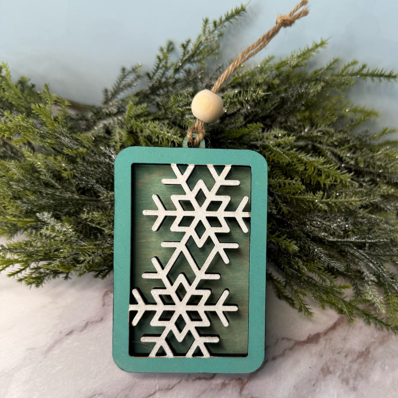 Decorative ornament with snowflake design on a teal background, hanging against a green leafy background.