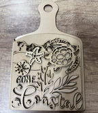 Wooden cutting board with coastal designs and text on a wooden surface