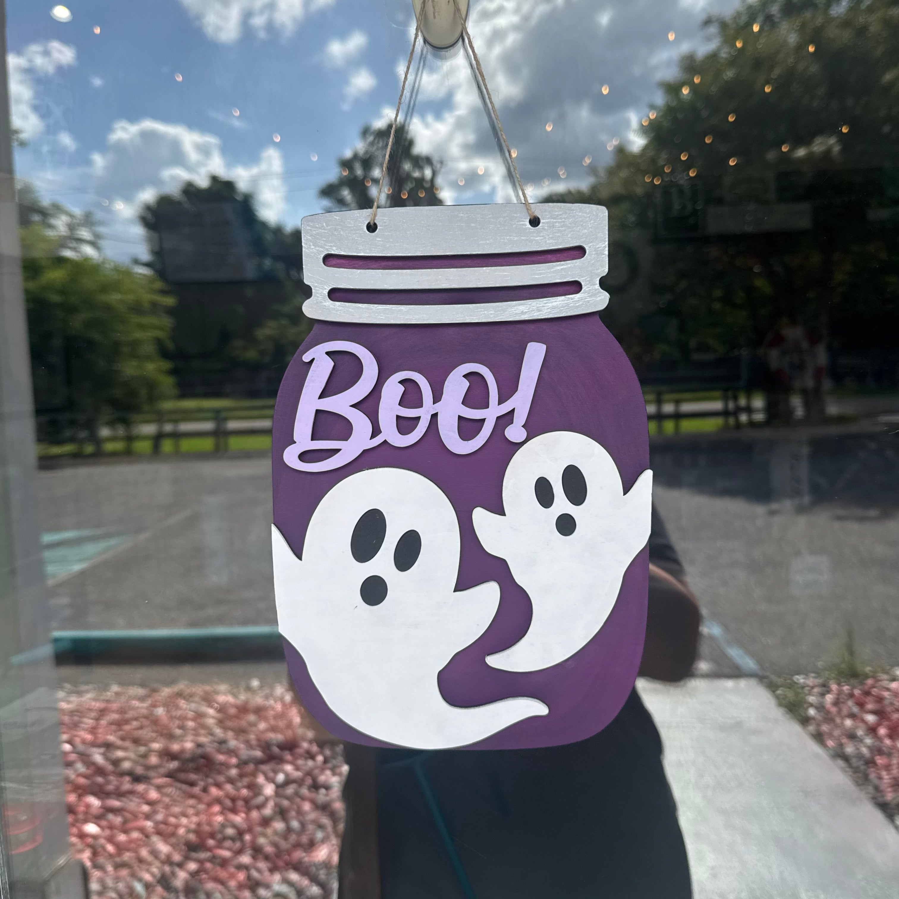 A purple mason-jar-shaped Halloween hanging decoration with white ghosts and the word "Boo!" in lavender, photographed hanging in front of a window with an outdoor scene behind it.