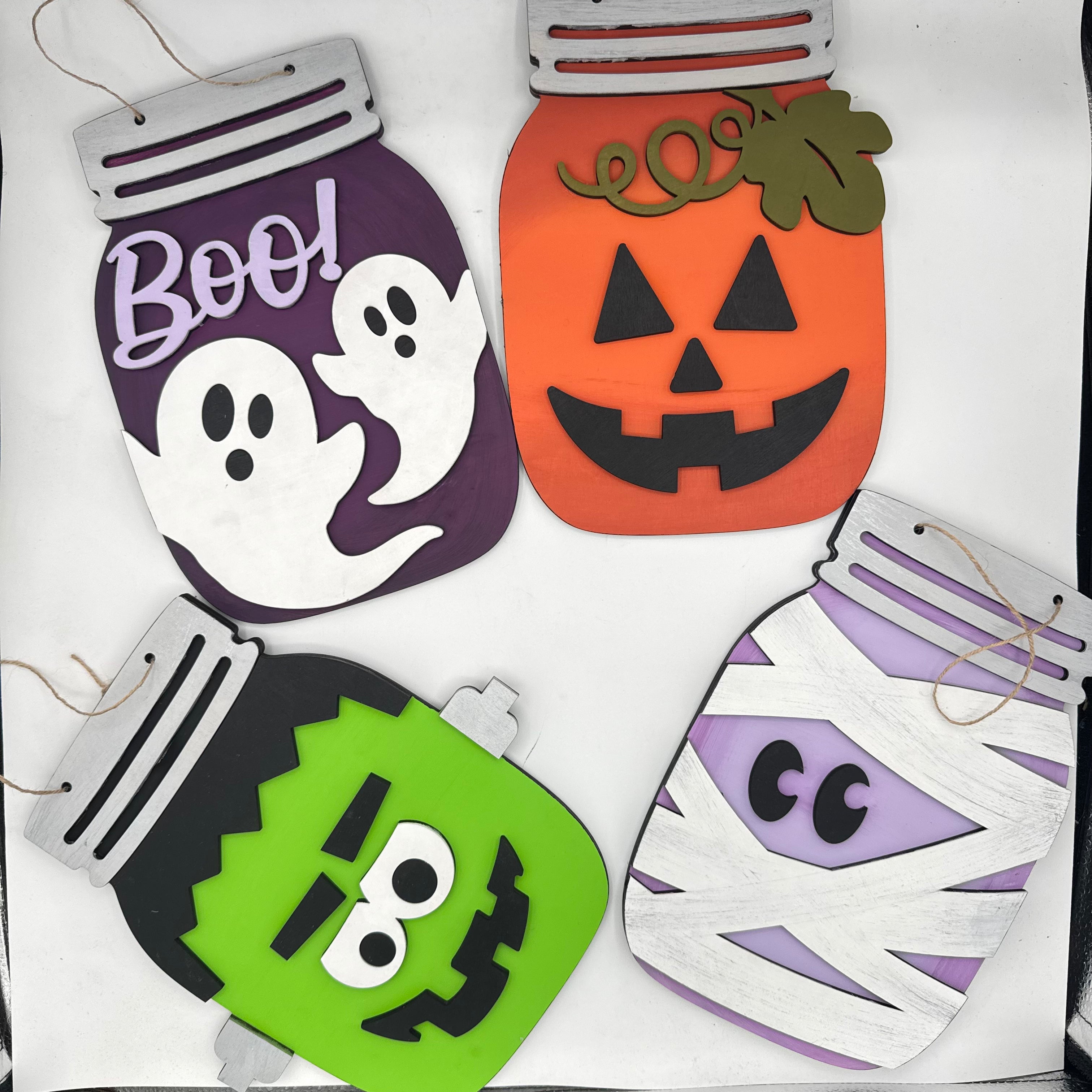 Four Halloween-themed jigsaw puzzle cutouts shaped like mason jars, arranged on a white background. The designs include a purple ghost jar with the word 'Boo!', an orange jack-o'-lantern jar, a green Frankenstein jar, and a white-and-purple mummy jar.