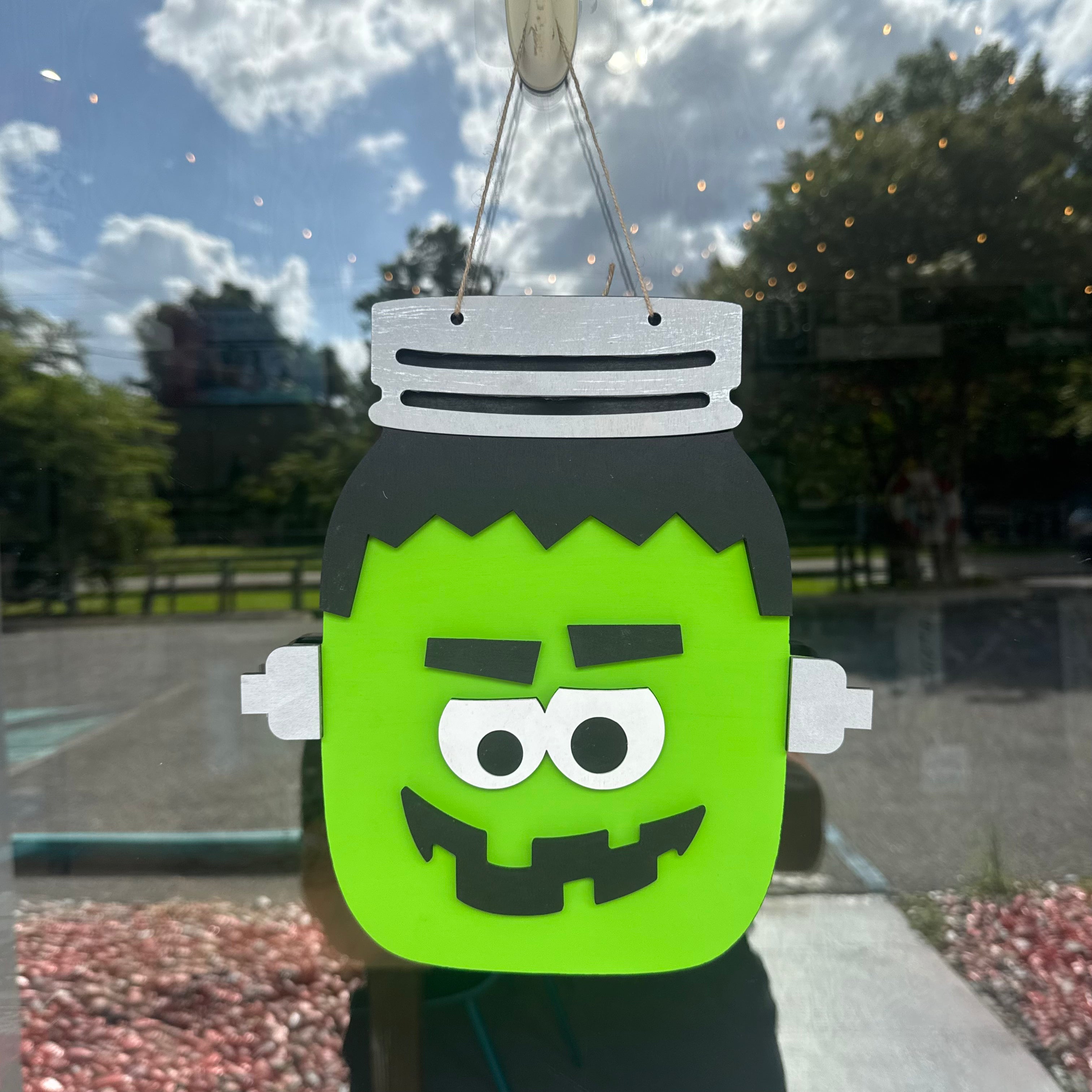 A Frankenstein-themed mason jar Halloween decoration hanging in front of a window. The decoration features bright green skin, black hair, white collar and hands, and a black stitched mouth and eyebrows, with a metal hanging loop at the top.
