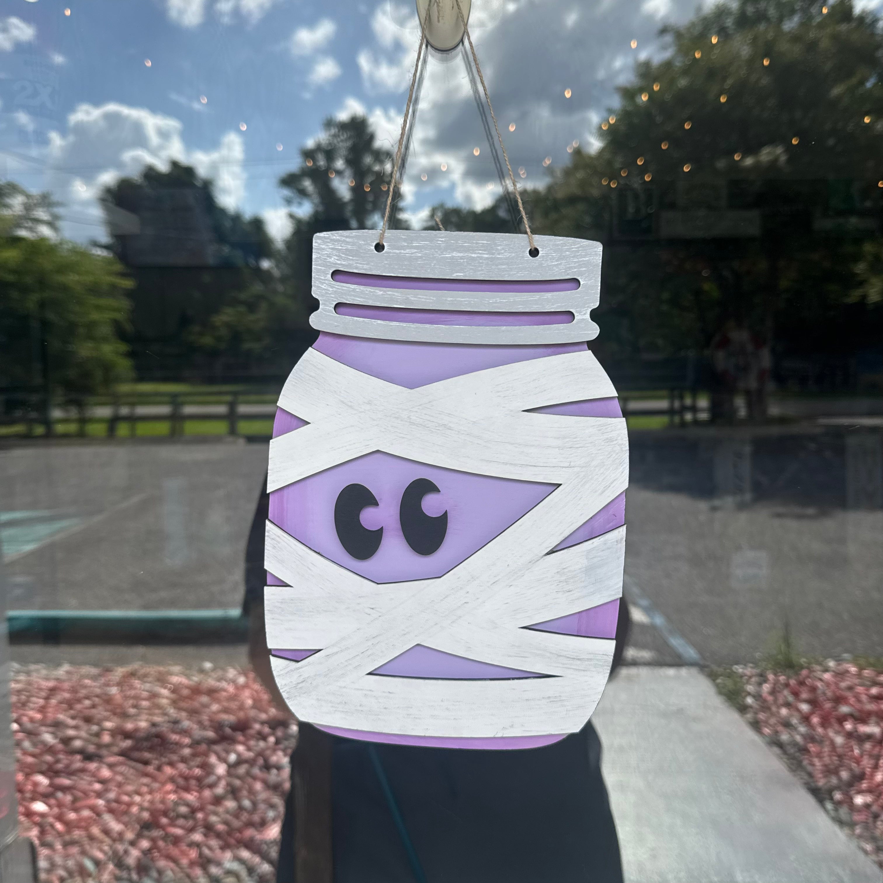 A wooden mason jar Halloween decoration wrapped in white wood-grain tape with a purple belly and black eyes, hanging in a window with an outdoor park background visible.