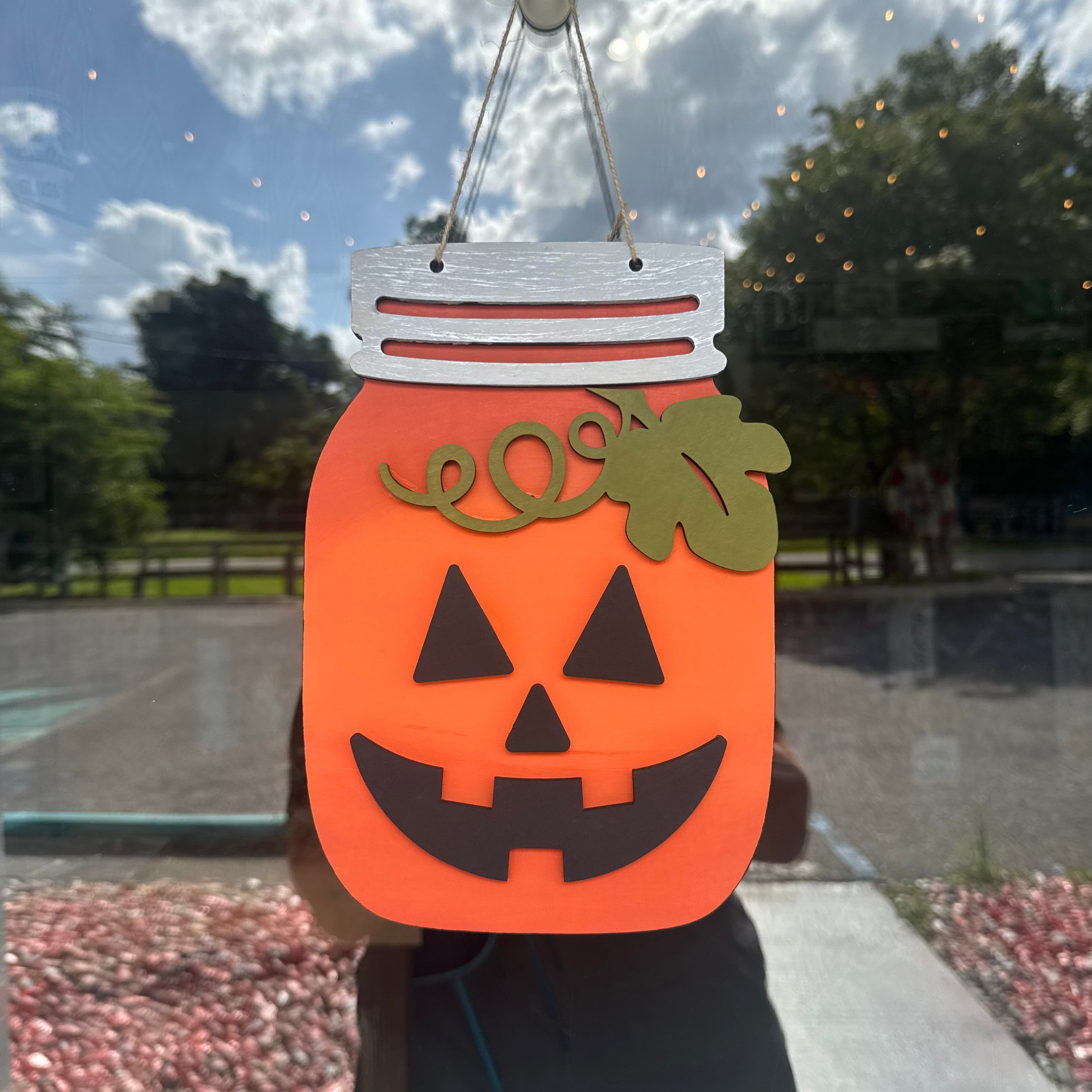 An orange jack-o'-lantern Halloween decoration shaped like a mason jar, hanging in front of a window. The decoration features a silver metal screw-top lid, a green leaf and curl accent on top, and a black face cutout.