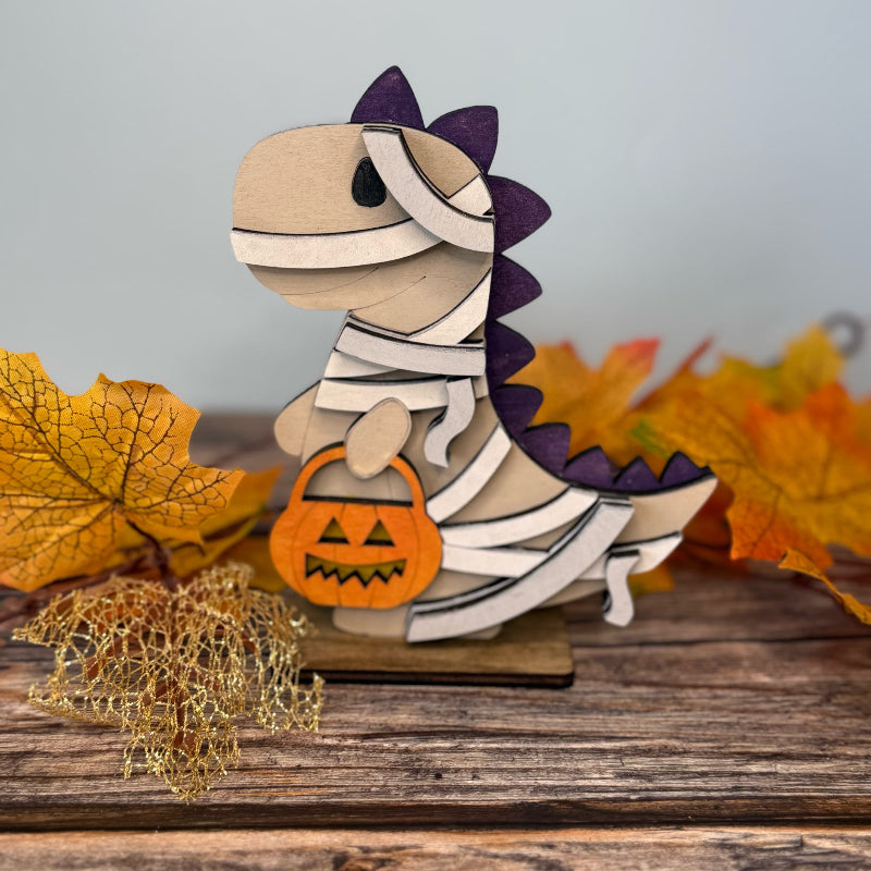 Wooden dinosaur mummy figurine with a pumpkin, surrounded by autumn leaves on a wooden surface.