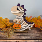 Wooden dinosaur mummy figurine with a pumpkin, surrounded by autumn leaves on a wooden surface.