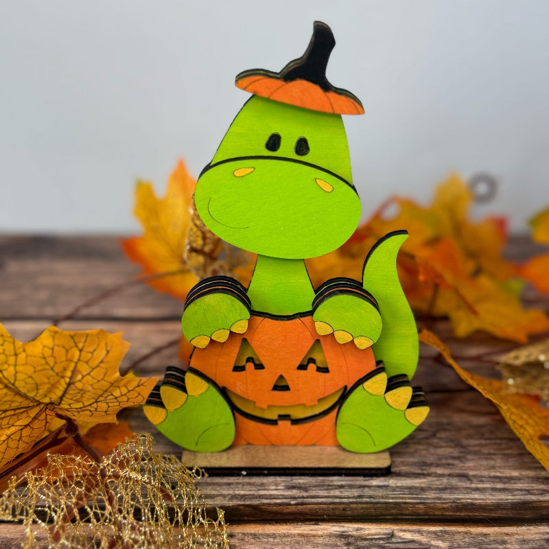 Green dinosaur with pumpkin hat and jack-o'-lantern belly on a wooden surface with autumn leaves.
