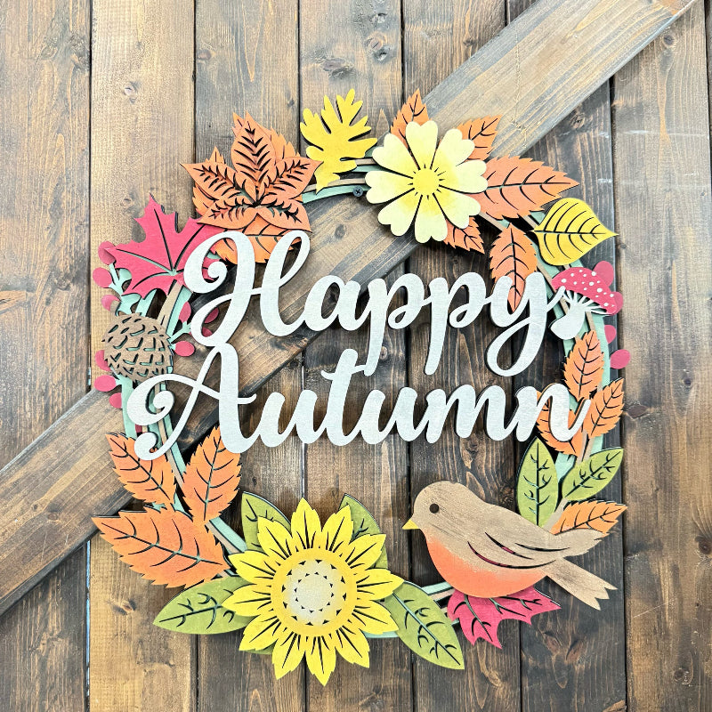 A decorative autumn wreath sign with layered paper cutout leaves, flowers, a bird, and the words “Happy Autumn” in white script, displayed against a rustic wooden plank background.